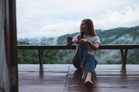 Portrait image of a woman drinking hot coffee while sitting on wooden balcony with a beautiful mountains and nature view on foggy day