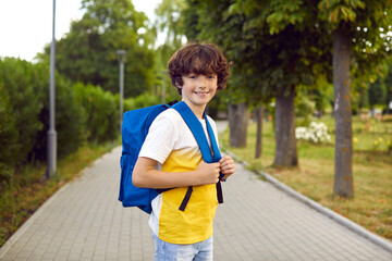 Portrait of cute preteen boy with school bag posing on warm morning on park walkway. Handsome caucasian little schoolboy with freckles wearing jeans and t-shirt smiling looking at camera.