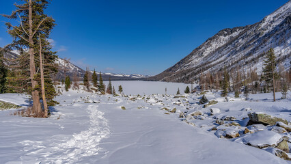 Fototapeta na wymiar Picturesque boulders are scattered in the snow-covered valley, coniferous trees grow. The path is trampled through snowdrifts. Ahead is a frozen lake surrounded by mountains. Altai. Multinskoye lake
