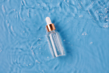 Glass bottle of anti aging serum and falling water drops on blue background