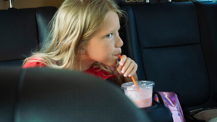 Beautiful Caucasian blonde girl drinks smoothies through a straw while traveling by car.