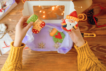 Women's hands in knitted sweater hold Christmas gingerbread with icing. Cookies in form of sock and Santa Claus. Gift box with paper, scissors, decorations and cones on wooden table. New Year mood.