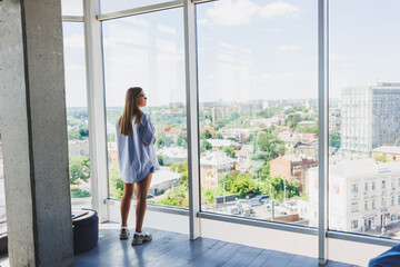 A young woman freelancer in glasses stands with coffee and talks on the phone near a large window and looks at the city. A girl in a shirt and shorts stands in a cafe by the window