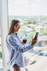 Happy woman drinks coffee and looks out the window in a cafe. A young smiling girl in glasses is standing by the window. Recreation, leisure and free time. The modern freelancer lifestyle
