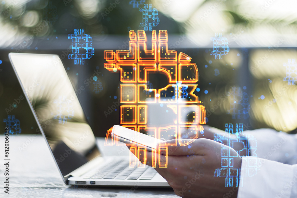 Poster hands using laptop and cellphone with abstract glowing bitcoin sign hologram on desktop and blurry outside background. Cryptocurrency, finance, trade and market concept. Double exposure.