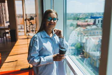 A young female freelancer in glasses is standing with coffee near a large window and looking at the city. A girl in a shirt and shorts stands in a cafe by the window