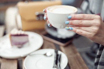 Close up of cup of cappuccino in female hands with blue manicure. Coffee shop outdoor.