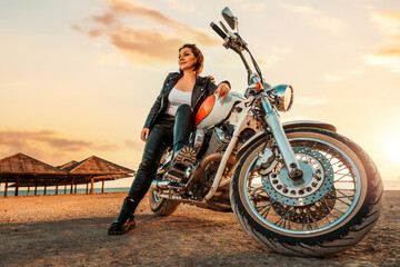 A beautiful independent woman in a leather clothes sitting on a motorcycle. Bottom view. Epic sunset on beach at the background. The concept of motorcycle trip and feminism