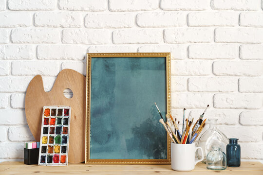 Photo frame as easel with artist's tools on wooden table against white brick wall