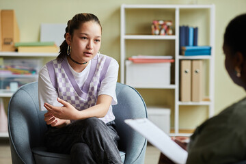 Teenage girl sitting on armchair and talking about her problems to psychologist during consultation...