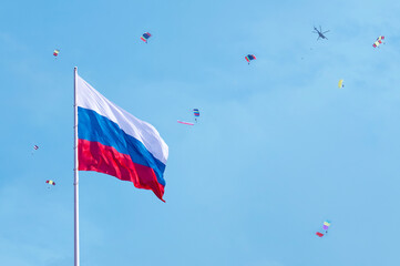 The national flag of the Russian Federation against the blue sky with a helicopter and paratroopers with colorful parachutes. Bright sunlight. Victory day celebration.