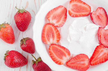 top view of yogurt with strawberry slices in white plate and whole strawberries on white wooden background