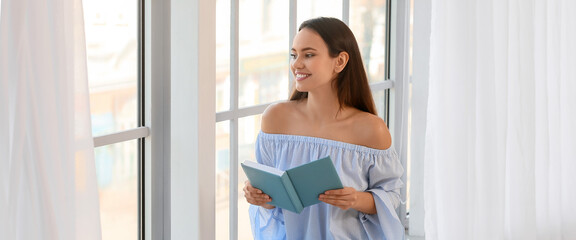 Pretty young woman reading book while sitting on window sill
