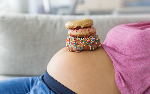 Pregnant woman with donuts on belly. Cravings of desserts and sweets during pregnancy, Girl eating unhealthy pastries on baby bump for gestational diabetes