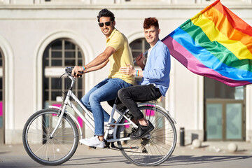 Side view of Young gay couple holding a gay pride flag while riding a bike outdoors. Concept of...