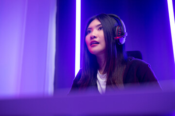 Pro gamer. Young pretty asian woman sitting on chair playing computer pc game online in the dark room with neon light. Chinese Teenage female with headphone professional gamer