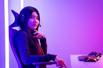 Fototapeta na wymiar Professional gamer. Young asian pretty woman sitting on chair with computer pc in living room. Happy female Professional Streamer chinese wearing headphone playing game online in dark room neon light