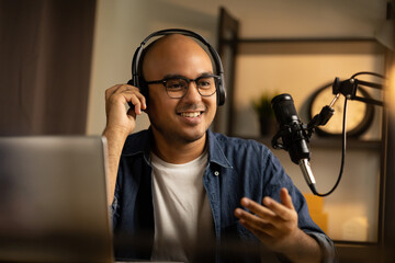 Young asian man host streaming podcast with condenser microphone work on laptop at small broadcast...