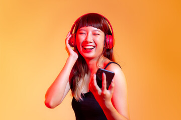Young asian woman in black tank top wearing red headphones and holding smartphone chatting and listen to music online on the orange background.