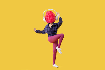 Woman with headphones and ripe raspberry instead of her head doing aerobics on yellow background - obrazy, fototapety, plakaty