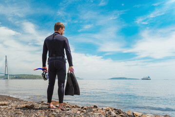 a diver in a wetsuit stands on the seashore. Diver before diving into the ocean, in the hands of a mask and fins for diving © ProstoSvet