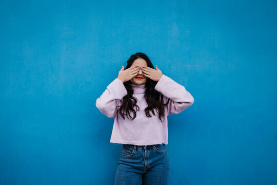 Young woman covering eyes with hands standing in front of blue wall