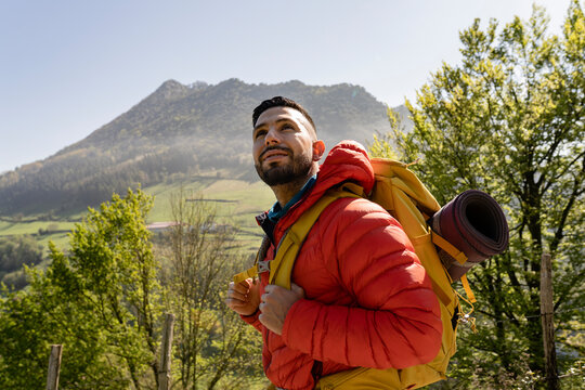 Smiling hiker wearing backpack standing in front of tree on sunny day