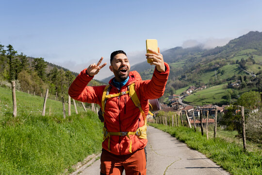 Happy man gesturing peace sign taking selfie through smart phone on sunny day