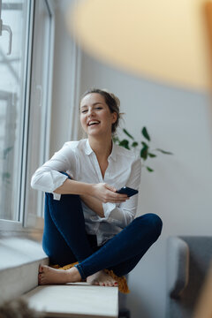 Happy woman holding smart phone sitting on widow sill at home