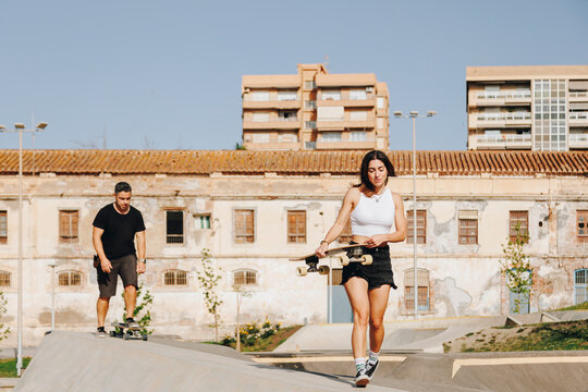 Young woman holding skateboard walking on sports ramp ahead of man