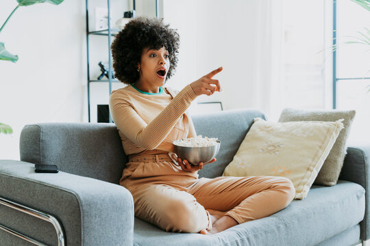 Shocked Afro woman watching TV sitting on sofa with bowl of popcorn pointing at home
