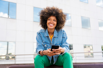 Beautiful happy woman with Afro hairstyle holding mobile phone sitting on bench