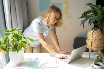 Mature freelancer wearing eyeglasses using laptop standing at desk in home office