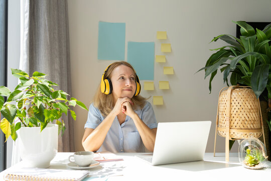 Thoughtful Smiling Freelancer Listening Music Through Wireless Headphones Sitting With Laptop At Desk In Home Office