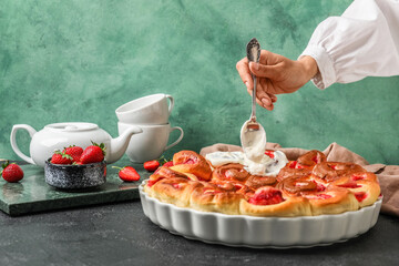 Female chef adding cream onto strawberry cinnamon rolls on table against green background