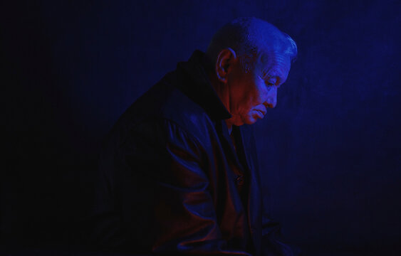 Depressed lonely senior man illuminated with blue neon light against black background