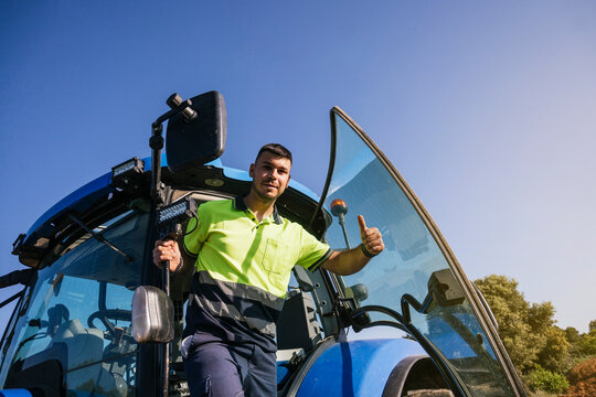 Young farmer gesturing thumbs up standing at door of tractor