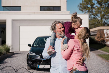 Woman kissing daughter sitting on man's shoulder in front of electric car
