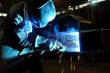 A welder in protective overalls and a welding mask welds metal structures by welding on a...