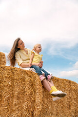 Young beautiful mother with little daughter sits on haystack. Happy childhood in the country. Summer vacation