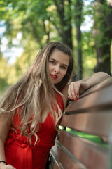 Sexy young woman in red dress with long hair sitting on bench in park. Attractive girl. Vertical frame