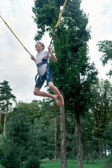 Happy laughing boy hangs on slings, jumps high on trampoline in an amusement park. Teenager is having fun.