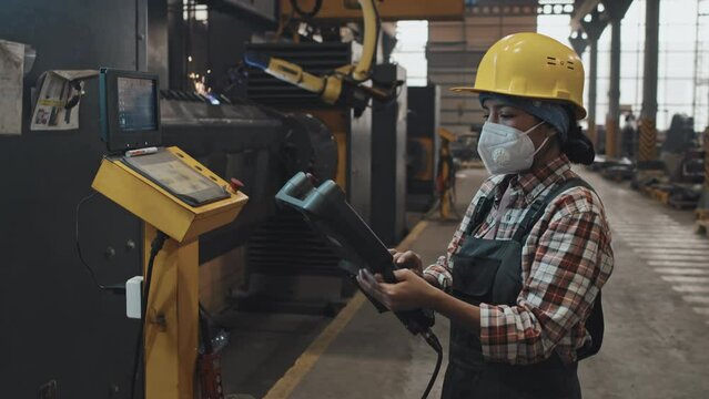 Medium side view of female Hispanic technician in yellow helmet, respirator and uniform operating special equipment at plant