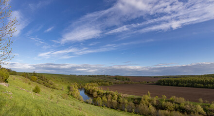 Landscape with a small river in a ravine. View of the countryside. bright greenery in the ravine. Saturated green grass against the blue sky. Plowed field on the horizon.