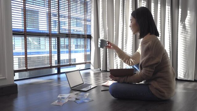 Freelance Asian woman sitting on floor while working and checking the best images from photoshoot and writing notes in notebook. Blogger, creative work online marketing. Startup Small business owner.