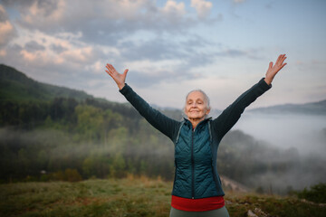Senior woman doing breathing exercise in nature on early morning with fog and mountains in...