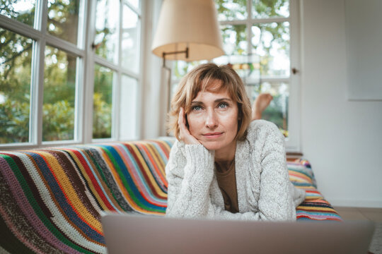 Thoughtful mature woman with laptop lying on sofa at home