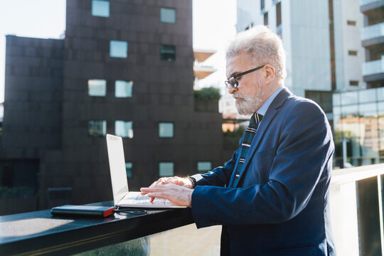 Senior Businessman Using Laptop By Solar Battery Charger Standing At Office Balcony