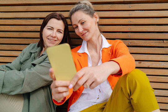 Smiling woman taking selfie with friend through mobile phone sitting on bench