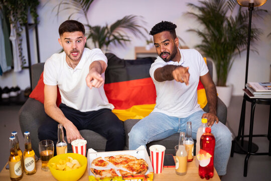 Young men pointing watching soccer match together sitting on sofa at home